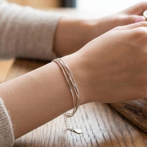 May include: A delicate silver bracelet with three strands is shown on a wrist. The bracelet has a simple design with a small, adjustable clasp and a teardrop-shaped charm. The jewelry is shown against a wooden surface.