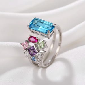 Può includere: Un anello argentato con una grande gemma rettangolare azzurra e un gruppo di gemme colorate più piccole. Il design dell'anello include pietre rosa, viola, verdi e blu, con una fascia ornata da piccoli cristalli trasparenti.