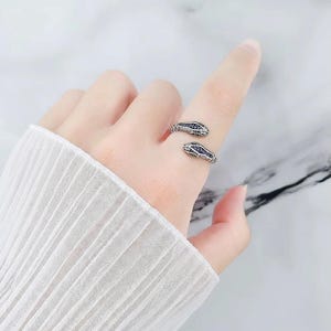 Könnte beinhalten: Ein silberner Schlangenring mit zwei Schlangenköpfen, die jeweils mit kleinen blauen und klaren Steinen verziert sind. Der Ring umschlingt den Finger und schafft ein einzigartiges und stilvolles Accessoire. Der Ring befindet sich an einer Hand mit einem weißen Ärmel.