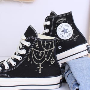 May include: Black high-top sneakers with white laces and soles. The sneakers feature decorative chain and cross designs, with the Converse All Star logo on the side. A pair of jeans is visible in the background.