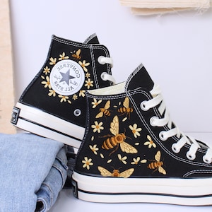 Puede incluir: Zapatillas altas negras con cordones y suelas blancas. Las zapatillas presentan un diseño de abejas y flores. El logotipo de Converse es visible en el lateral. Los zapatos están colocados sobre una superficie blanca.