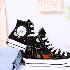 May include: Black high-top sneakers with Halloween-themed embroidery. The design includes ghosts, a haunted house, pumpkins, and fall foliage. The sneakers have white laces and soles, with a classic Converse logo on the side.