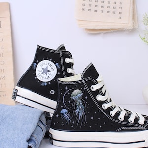 Puede incluir: Zapatillas altas negras con cordones y suelas blancas. Las zapatillas presentan un logotipo de estrella y un diseño de medusa. Se combinan con un par de vaqueros azules doblados.