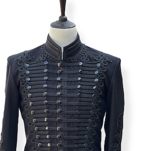Può includere: Una giacca nera in stile militare con colletto alto e dettagli intricati in treccia nera. La giacca ha una fila di bottoni sul davanti e un design decorativo a treccia sulle maniche.