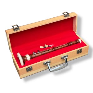 Puede incluir: Una flauta dulce de madera en un estuche marrón claro con interior de terciopelo rojo. La flauta dulce es de color marrón oscuro con detalles blancos y crema. El estuche tiene cierres y un asa plateados. Se incluyen piezas adicionales de flauta dulce.