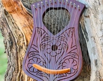May include: A wooden lyre with a floral design carved into the body. The lyre has 15 strings and a wooden bridge. The lyre is resting on a tree trunk.
