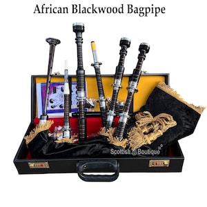 Puede incluir: Gaitas de ébano africano exhibidas en un estuche negro con interior rojo. Las gaitas son negras con detalles plateados y borlas doradas. El estuche tiene un asa y cerraduras de combinación. El texto "Scottish Boutique" es visible.