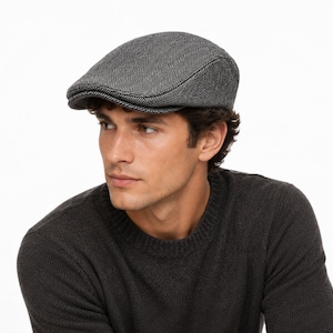 May include: A grey and black flat cap featuring a classic herringbone pattern. The cap has a structured crown and a short, stiff brim that extends forward. The fabric displays a woven texture with alternating light and dark grey threads.