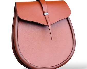 May include: A brown leather bag with a rounded bottom and a flap closure. The bag has a strap that secures the flap with a silver-colored metal detail. The bag is designed with a textured surface and a smooth, curved trim.