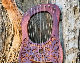 May include: A wooden lyre with intricate floral carvings and multiple strings. The lyre is brown with a reddish hue and is resting against a tree trunk.