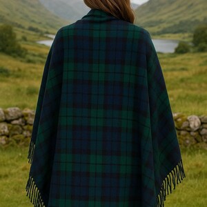 May include: A dark green and navy blue plaid cape with fringe trim. The cape is worn outdoors with a scenic background of mountains and a lake. The plaid pattern is a classic design, suitable for cool weather.