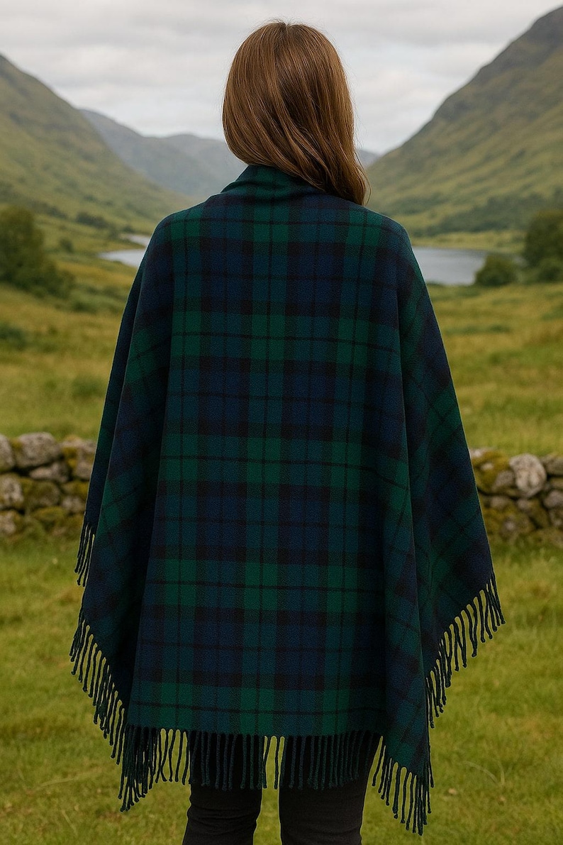 May include: A dark green and navy blue plaid cape with fringe trim. The cape is worn outdoors with a scenic background of mountains and a lake. The plaid pattern is a classic design, suitable for cool weather.