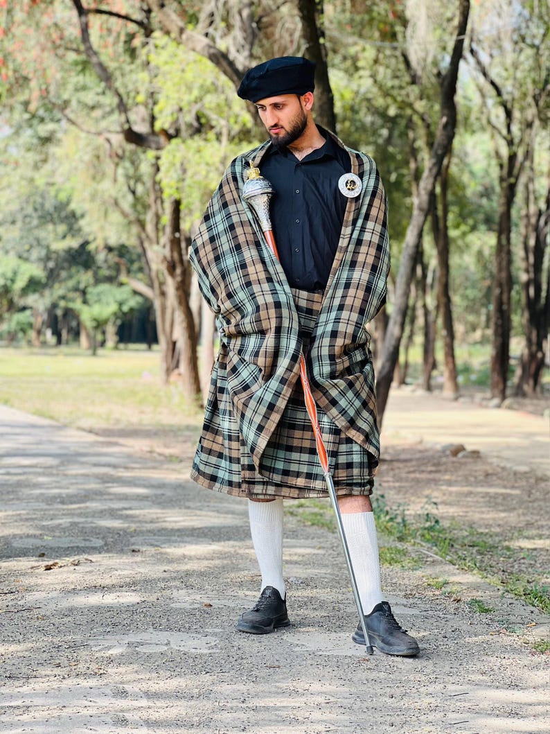Peut inclure: Un b&eacute;ret noir, une chemise noire boutonn&eacute;e et un kilt &agrave; carreaux dans des tons de beige, vert et noir. La tenue comprend un ch&acirc;le &agrave; carreaux assorti drap&eacute; sur les &eacute;paules, des chaussettes blanches hautes, des baskets noires et une masse.