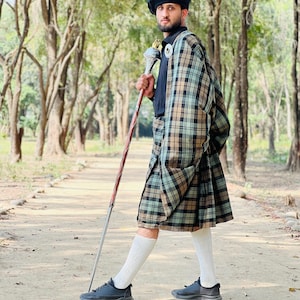 Peut inclure: Un b&eacute;ret noir, une chemise noire &agrave; col et un kilt &agrave; carreaux dans des tons de beige, marron et vert clair. La tenue comprend des chaussettes blanches arrivant aux genoux, des baskets noires et un long b&acirc;ton m&eacute;tallique fin avec un embout d&eacute;coratif argent&eacute; et dor&eacute;.