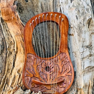 Può includere: Una lira di legno con un design floreale intagliato, appoggiata su un tronco d'albero. La lira ha 10 corde e un ponte di legno.