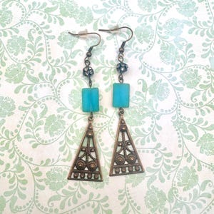 May include: A pair of earrings with turquoise blue rectangular beads, bronze-colored triangle pendants, and decorative accents. The earrings hang from fish hook ear wires. The background is a light green floral pattern.