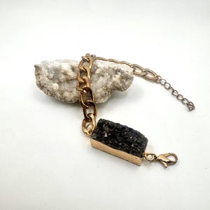 May include: A gold chain bracelet with a black rectangular stone pendant. The stone has a rough, textured surface.