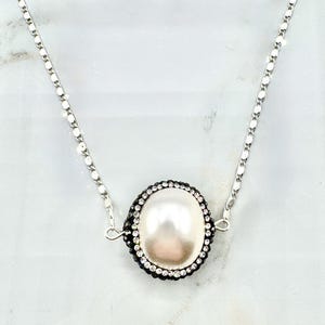 May include: A silver necklace featuring a large, oval, off-white pearl pendant. The pearl is encircled by a band of small, black and clear crystals. The chain is a delicate silver link design, creating a classic and elegant jewelry piece.