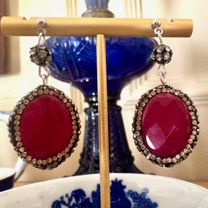 May include: A pair of dangle earrings with a large oval-shaped red gemstone surrounded by a halo of small clear rhinestones. The earrings have a small round rhinestone at the top of the dangle.