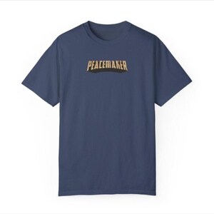 May include: A navy blue t-shirt with the word "PEACEMAKER" printed across the chest in a gold and black font. The shirt has short sleeves and a crew neck. The background is white.