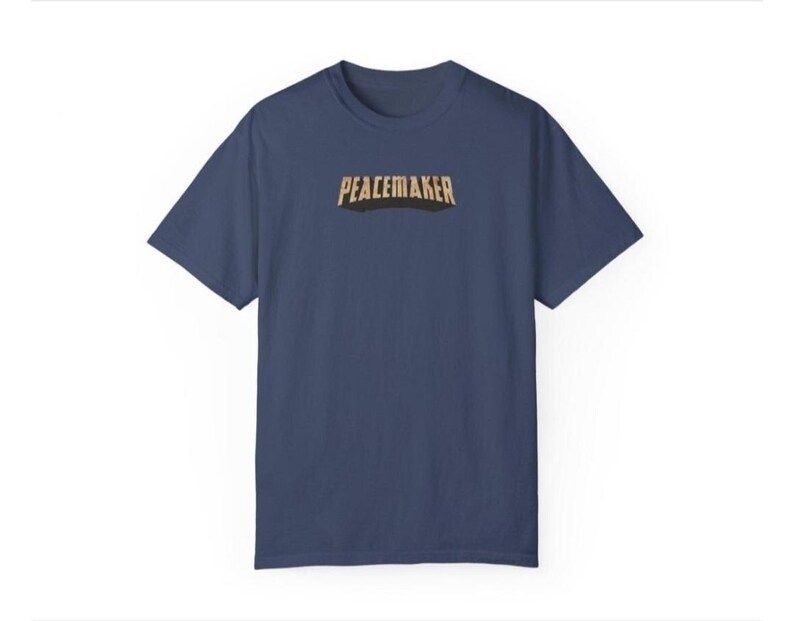 May include: A navy blue t-shirt with the word "PEACEMAKER" printed across the chest in a gold and black font. The shirt has short sleeves and a crew neck. The background is white.