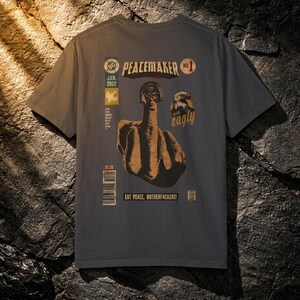 May include: Dark gray t-shirt with a comic book style graphic on the back. The graphic features a figure giving the middle finger, with text that reads "PEACEMAKER" and "EAT PEACE, MOTHERFUCKERS!"