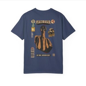 May include: Navy blue t-shirt with a comic book style graphic. The graphic features a figure giving the middle finger, with the text "PEACEMAKER" and "EAT PEACE, MOTHERFUCKERS!". Additional text includes "JAN 2022" and a barcode.