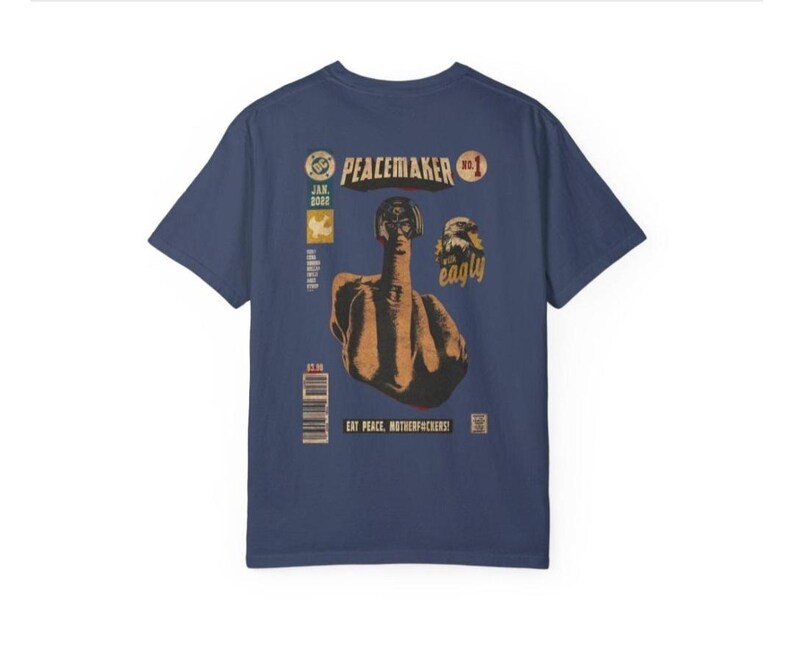 May include: Navy blue t-shirt with a comic book style graphic. The graphic features a figure giving the middle finger, with the text "PEACEMAKER" and "EAT PEACE, MOTHERFUCKERS!". Additional text includes "JAN 2022" and a barcode.