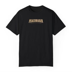 May include: Black t-shirt with the word "PEACEMAKER" in a gold, slightly arched font across the chest. The shirt has short sleeves and a crew neck. The design is centered on the front of the shirt.