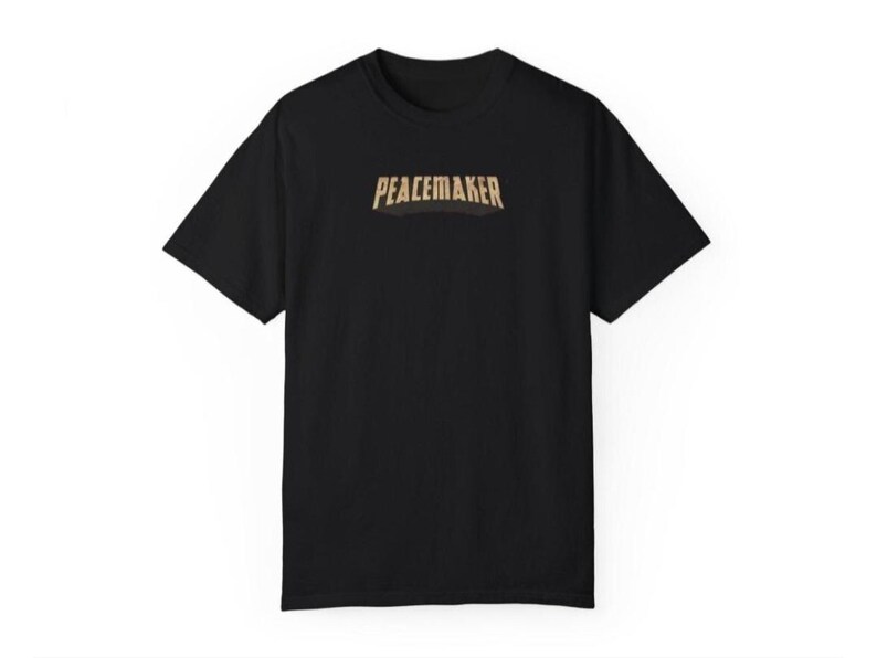 May include: Black t-shirt with the word "PEACEMAKER" in a gold, slightly arched font across the chest. The shirt has short sleeves and a crew neck. The design is centered on the front of the shirt.