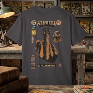 May include: Dark gray t-shirt featuring a comic book style design with the text "PEACEMAKER" and "EAT PEACE, MOTHERF#CKERS!". The graphic includes a figure with a raised middle finger and the word "eagly".