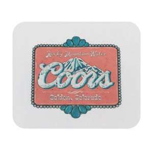 以下が含まれることがあります： Coorsのロゴと「Rocky Mountain Water」と「Golden, Colorado」の文字が入ったヴィンテージデザインの正方形コースター。デザインには山脈が含まれており、ピーチ色の背景にターコイズのアクセントとビーズの縁取りが施されています。