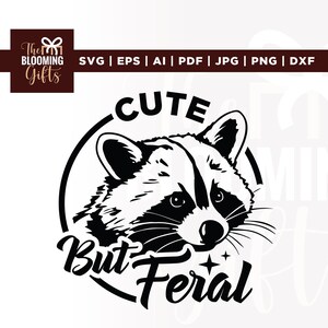 May include: Black and white graphic of a raccoon face inside a circle. The text above the raccoon reads "CUTE", and the text below reads "But Feral". The Blooming Gifts logo is in the upper left corner.