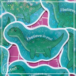 Könnte beinhalten: Ein gestanzter Aufkleber mit einem grünen langhalsigen Dinosaurier, der in Wasser mit Blasen schwimmt. Der Text "I believe in you!" ist in Weiß über die Mitte des Dinosauriers gedruckt. Der Aufkleber hat ein holografisches Finish mit kreisförmigen Mustern.