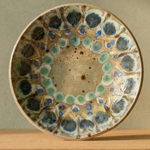 Può includere: Una ciotola in ceramica rotonda con un centro beige maculato circondato da un bordo decorativo di motivi smaltati sovrapposti in blu, verde e crema. Il bordo è liscio e circolare, mostrando una finitura a smalto reattivo con motivi circolari blu scuro e verde chiaro disposti in un design radiale attorno alla base interna.
