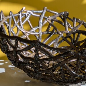 May include: A handcrafted, decorative bowl with a unique, openwork design. The bowl is made of dark, metallic-colored material, possibly metal or ceramic, with a woven, branch-like pattern. The bowl's interior is visible through the open design.