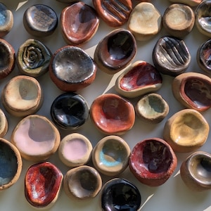Può includere: Una collezione di piccoli piatti in ceramica fatti a mano con forme rotonde e irregolari. Ogni pezzo presenta una finitura smaltata unica in vari colori, tra cui rossi profondi, neri lucidi, rosa tenui, marroni terrosi e tonalità crema.