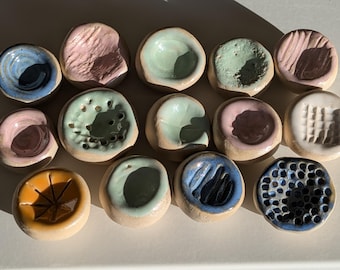 May include: A collection of fourteen small, round, handmade ceramic dishes with various glazed finishes. The pieces feature diverse textures, including smooth, pitted, and grooved surfaces in colors such as blue, pink, green, yellow, and white.