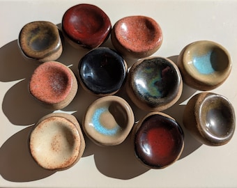 May include: A collection of small, handmade ceramic pinch pots in various colors. The pots have a rounded shape with a small, recessed interior. Colors include red, black, brown, and blue. The pots are arranged on a light-colored surface.
