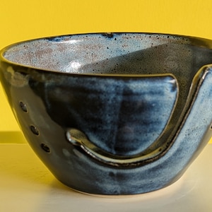 May include: A handmade ceramic yarn bowl with a glossy, dark blue glaze. The bowl features a curved slot for yarn and several small holes. The interior has a speckled, light blue and brown finish. The bowl is designed for knitting and crochet projects.