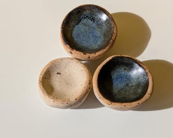May include: Three small, handmade ceramic bowls. Two bowls have a dark blue and black glaze, while the third is a speckled beige. The bowls have a rustic, organic feel, with visible texture and a natural, earthy aesthetic.