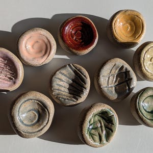 Può includere: Una collezione di dieci piccoli piatti in ceramica rotondi, formati a mano, con esterni in argilla maculata e testurizzata. Ogni pezzo presenta un centro smaltato unico in colori come rosa, rosso, giallo, beige, grigio e verde, con vari motivi.