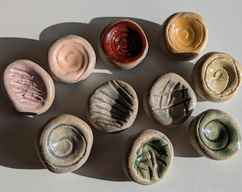 May include: A collection of ten small, round, hand-formed ceramic dishes with textured, speckled clay exteriors. Each piece features a unique glazed center in colors including pink, red, yellow, tan, gray, and green, with various swirl and line patterns.