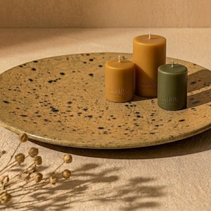 Op de afbeelding: Een ronde, gespikkelde beige keramische schaal met drie cilindrische stompkaarsen. Twee kaarsen zijn mosterdgeel en één is olijfgroen, elk voorzien van de inscriptie DARIUS. Een kleine, gedroogde botanische tak bevindt zich op de voorgrond.