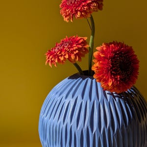 以下が含まれることがあります： 青いセラミックの花瓶には、テクスチャ加工された彫刻デザインがあり、鮮やかなオレンジ色のガーベラデイジーが3つ入っています。 花は濃い緑色の茎を持ち、黄色の背景に配置され、印象的なコントラストを生み出しています。