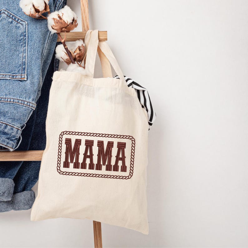 May include: Cream-colored tote bag with the word "MAMA" in a brown, Western-style font, framed by a brown rope design. The bag hangs on a wooden ladder with a pair of blue jeans and cotton stems.