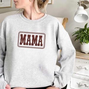 May include: A light gray sweatshirt with the word "MAMA" embroidered in a brown, Western-style font. The text is framed by a brown rope-like border. The sweatshirt has long sleeves.