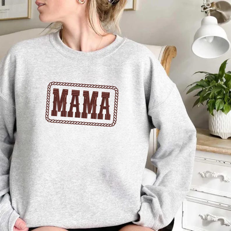 May include: A light gray sweatshirt with the word "MAMA" embroidered in a brown, Western-style font. The text is framed by a brown rope-like border. The sweatshirt has long sleeves.
