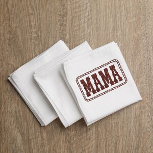 May include: Three white cloth napkins, one with the word "MAMA" embroidered in brown within a rope-style border. The napkins are stacked on a wooden surface.