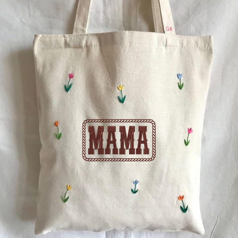 May include: Cream-colored tote bag with embroidered floral accents and the word "MAMA" in a brown, rope-framed design. The bag features small, colorful embroidered flowers scattered across the front, with a matching cream-colored handle.
